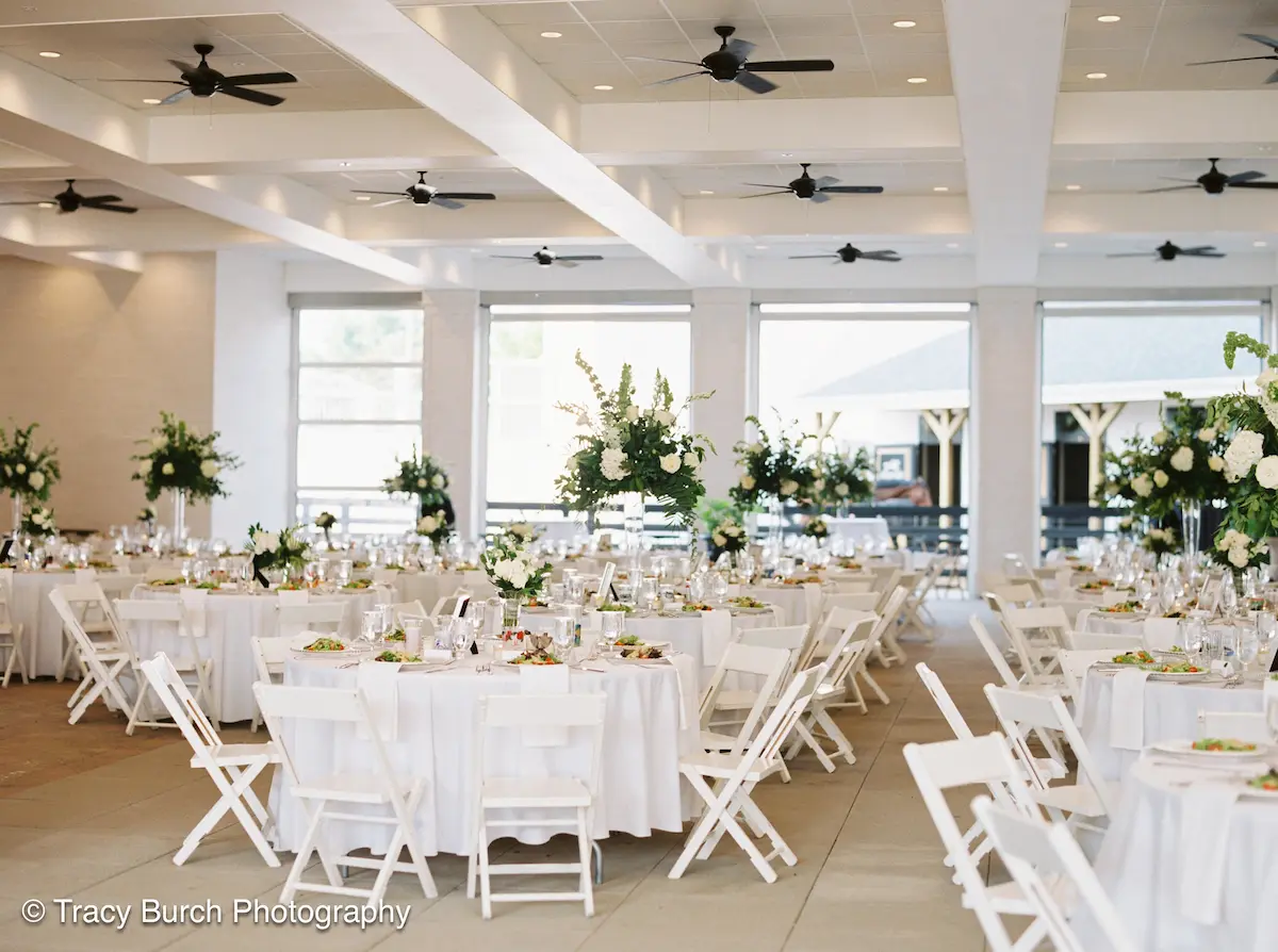 Spacious reception hall with round tables covered in white tablecloths, white folding chairs, and floral centerpieces.