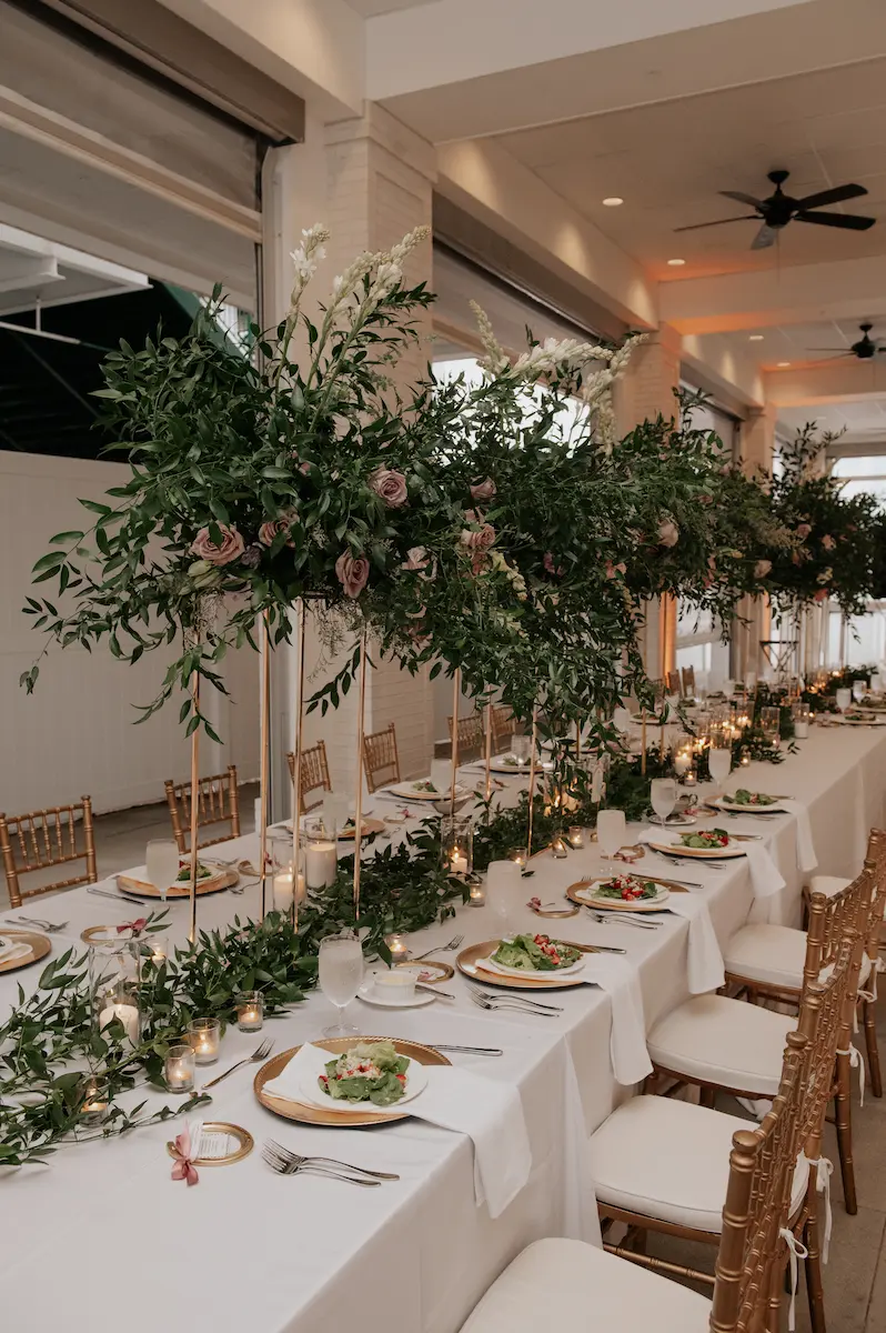 Elegant long dining table with white tablecloth, gold chairs, tall floral centerpieces with greenery and pink flowers, and candles in clear holders.