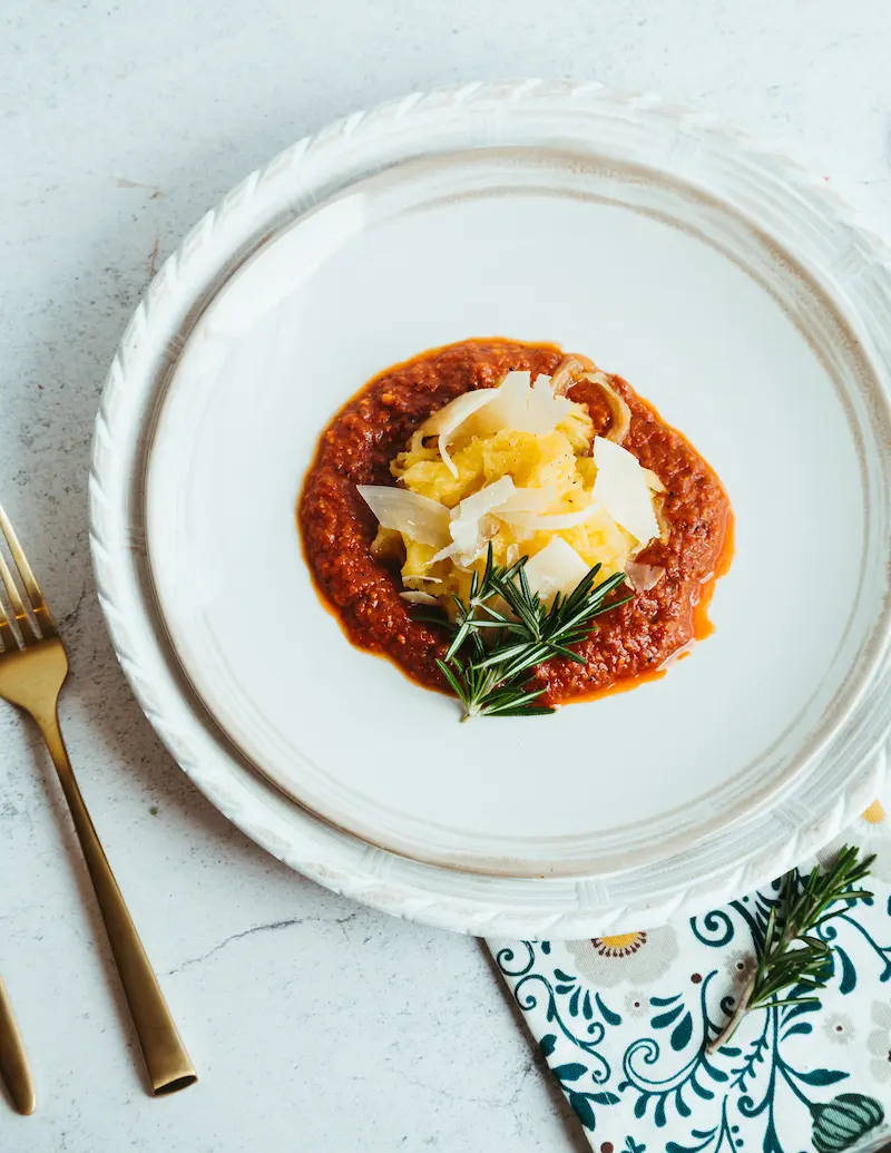 A serving of spaghetti squash with tomato sauce, shaved cheese, and a sprig of rosemary on a white plate with gold cutlery and a floral napkin nearby.