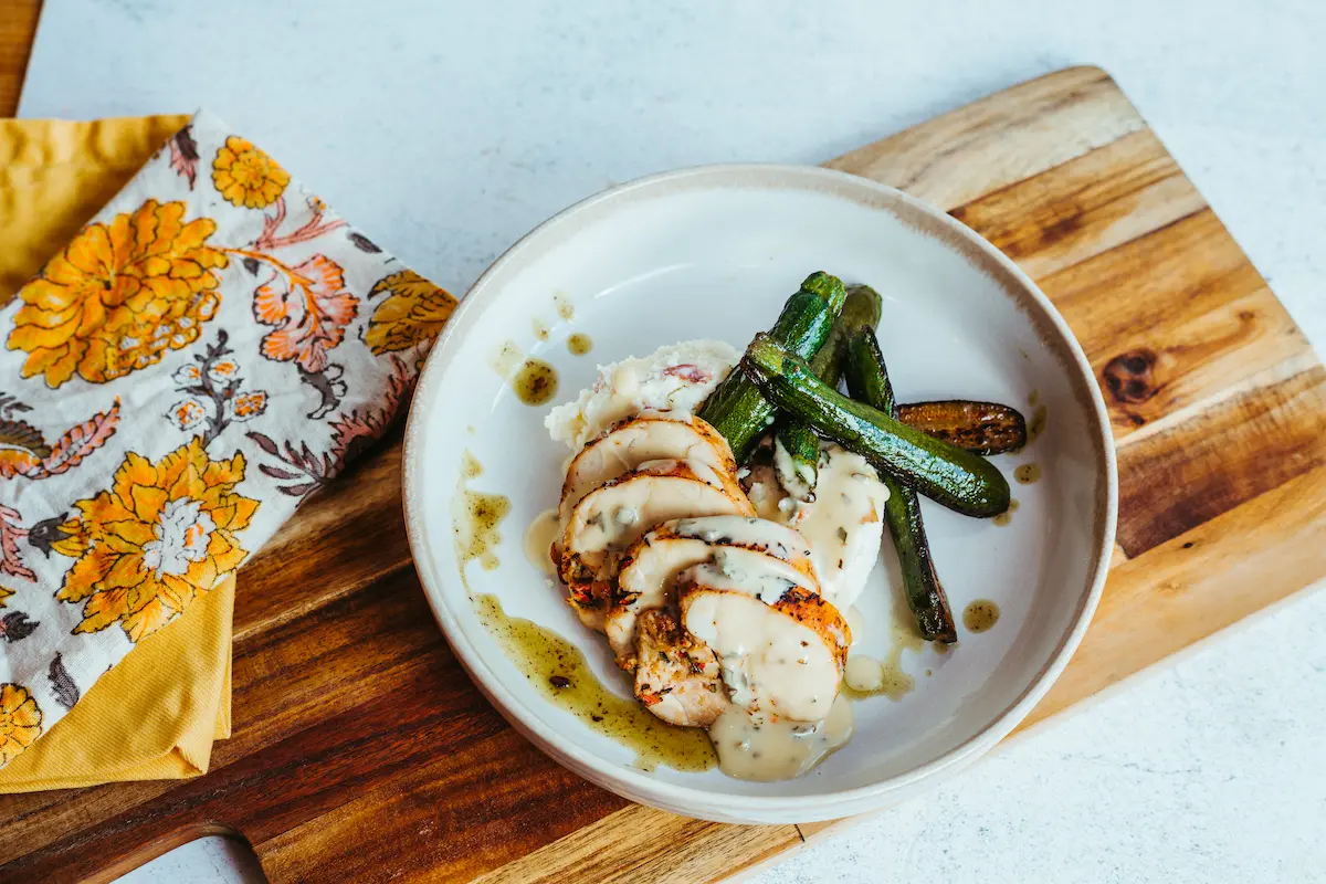 Plate with sliced grilled chicken breast topped with creamy sauce, small roasted zucchini, and mashed potatoes on a wooden cutting board with yellow floral napkins nearby.