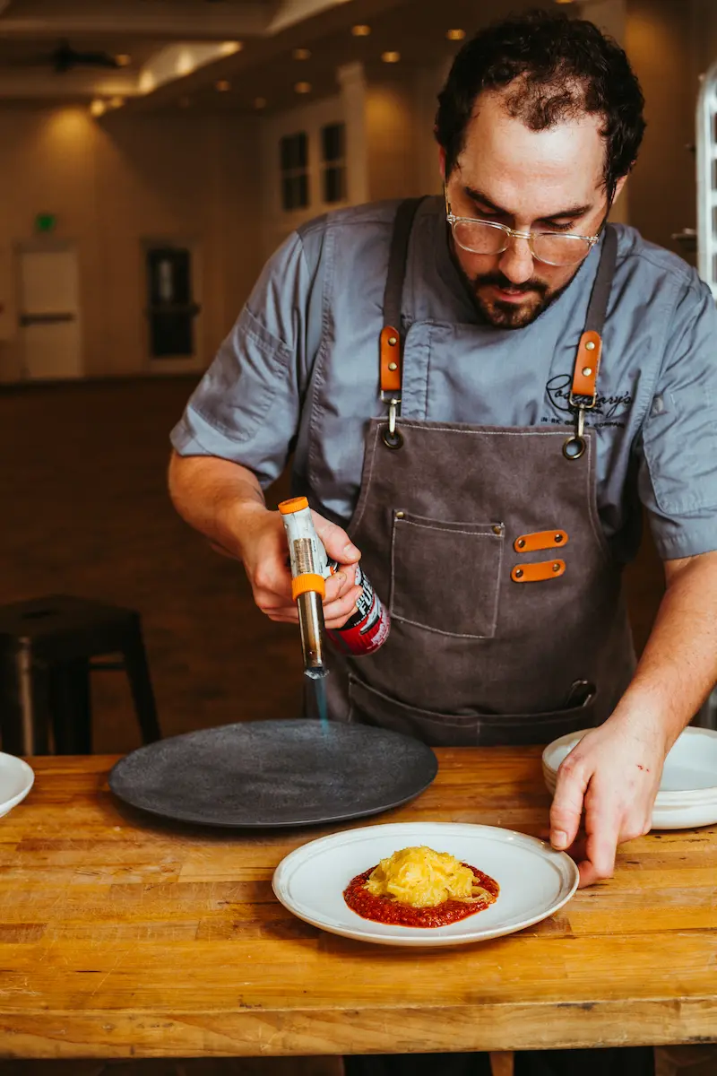 Chef using a blowtorch on a black plate beside a dish of spaghetti squash with tomato sauce on a wooden table.