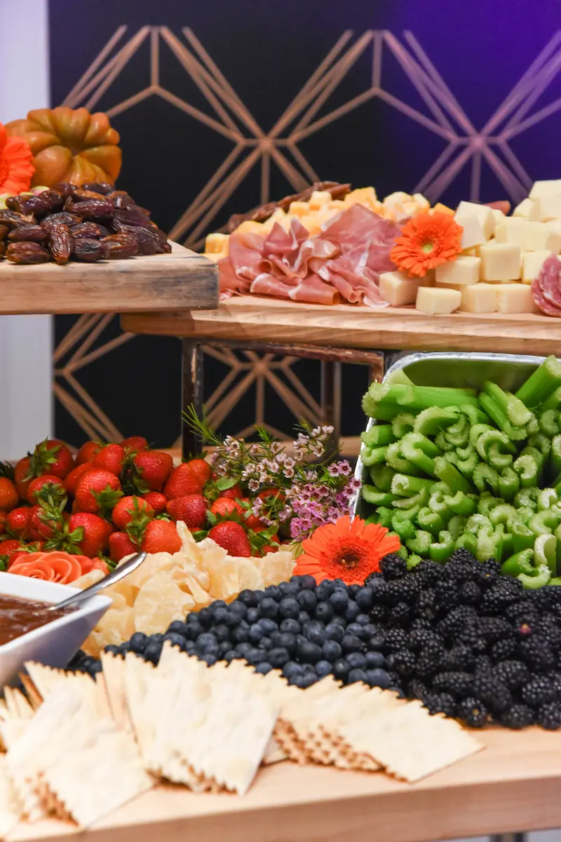 Charcuterie board with crackers, blueberries, blackberries, strawberries, celery, sliced meats, cubed cheeses, dates, and garnished with orange flowers.
