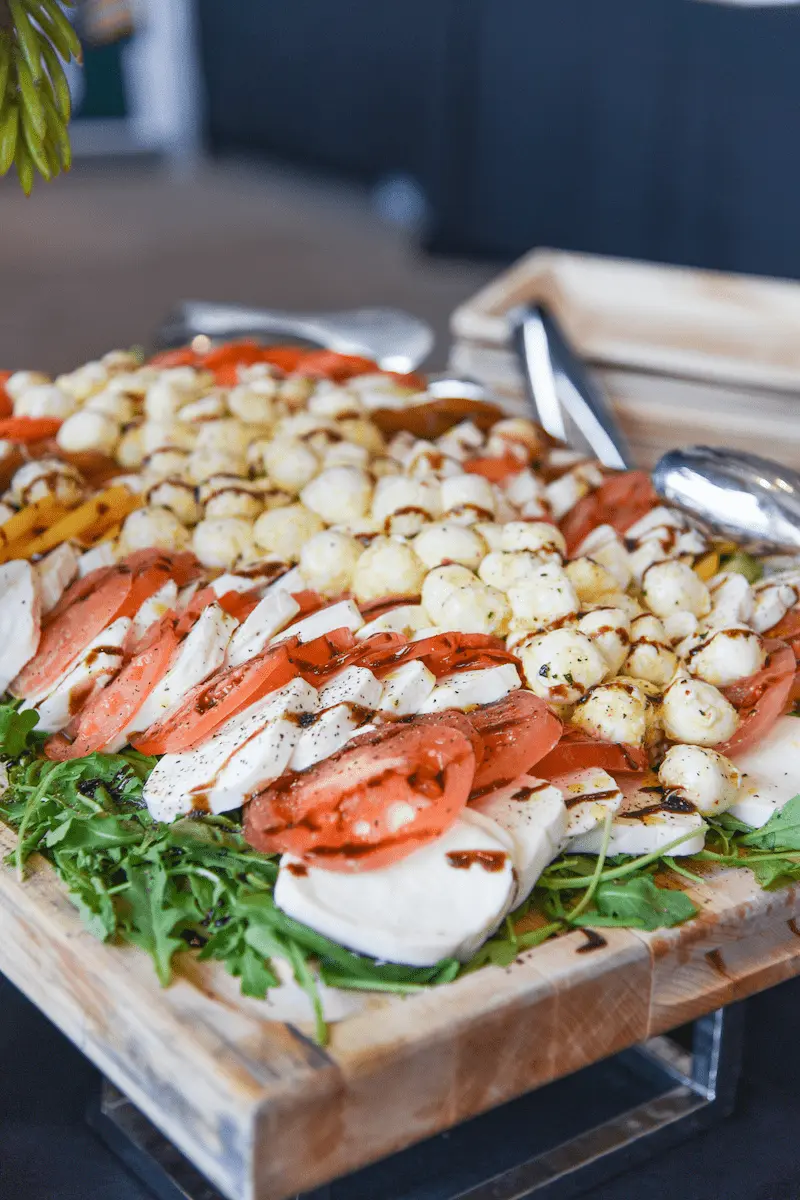Wooden tray with sliced tomatoes, mozzarella balls, fresh mozzarella slices, and arugula drizzled with balsamic glaze.