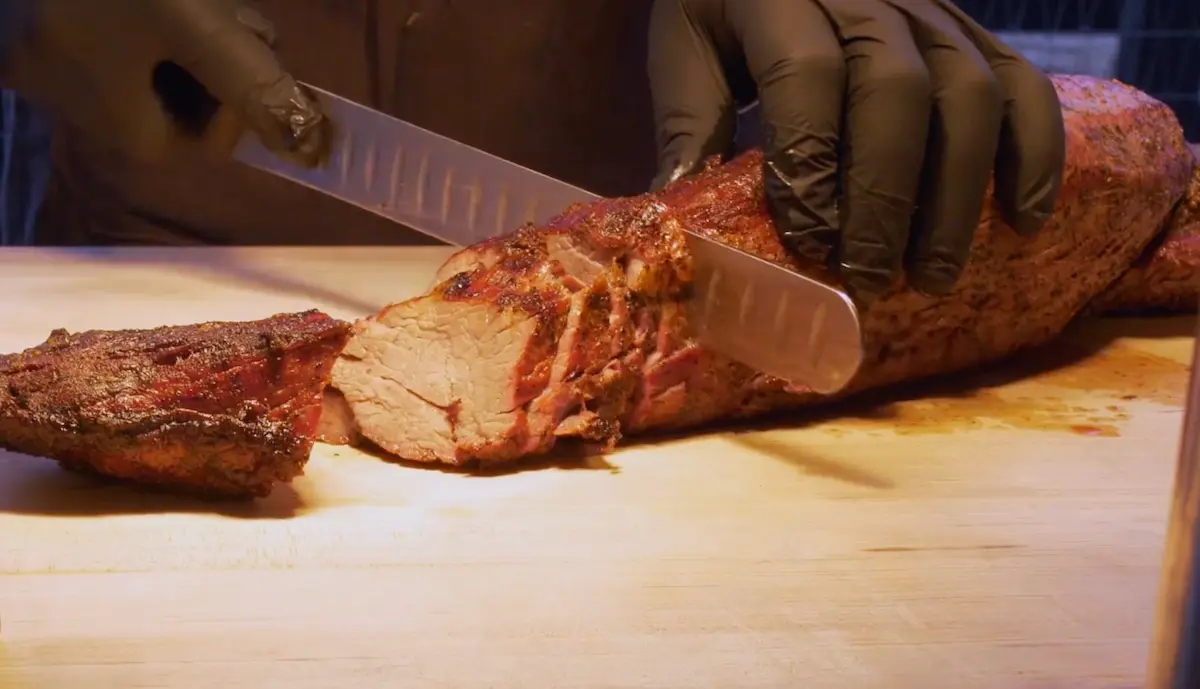 Person wearing black gloves slicing a cooked, seasoned piece of meat on a wooden cutting board.