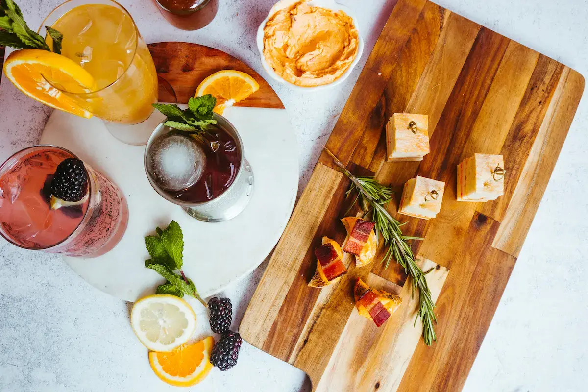 Wooden board with rosemary, small cheese cubes, and crackers topped with meat.