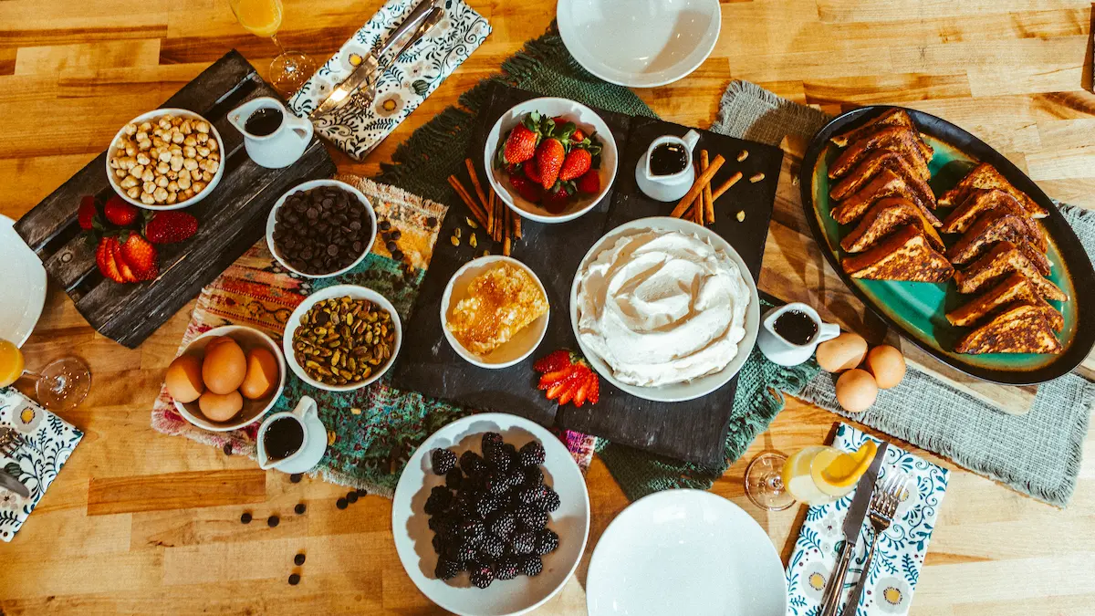 Table set with plates of whipped cream, blackberries, strawberries, toasted sandwiches, eggs, nuts, chocolate chips, and syrup pitchers.