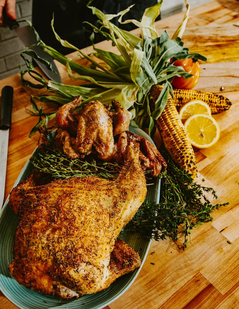 Roasted whole chicken and Cornish hens seasoned with herbs on a platter, accompanied by grilled corn, lemon slices, and fresh herbs on a wooden table.