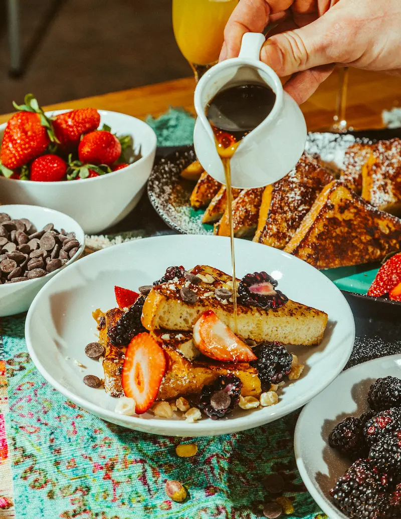 Hand pouring syrup over French toast topped with strawberries, blackberries, chocolate chips, and nuts on a white plate.