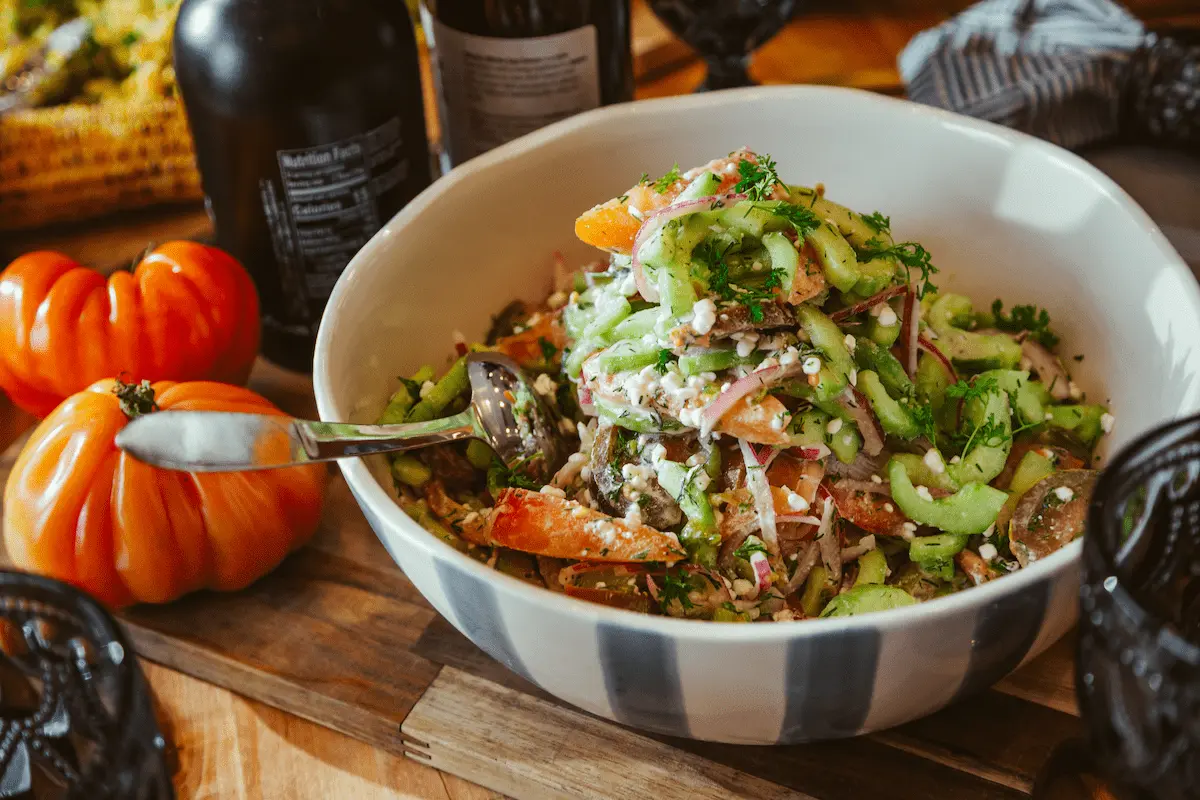 Bowl of fresh salad with celery, red onions, herbs, and cottage cheese on a wooden board next to tomatoes and bottles.