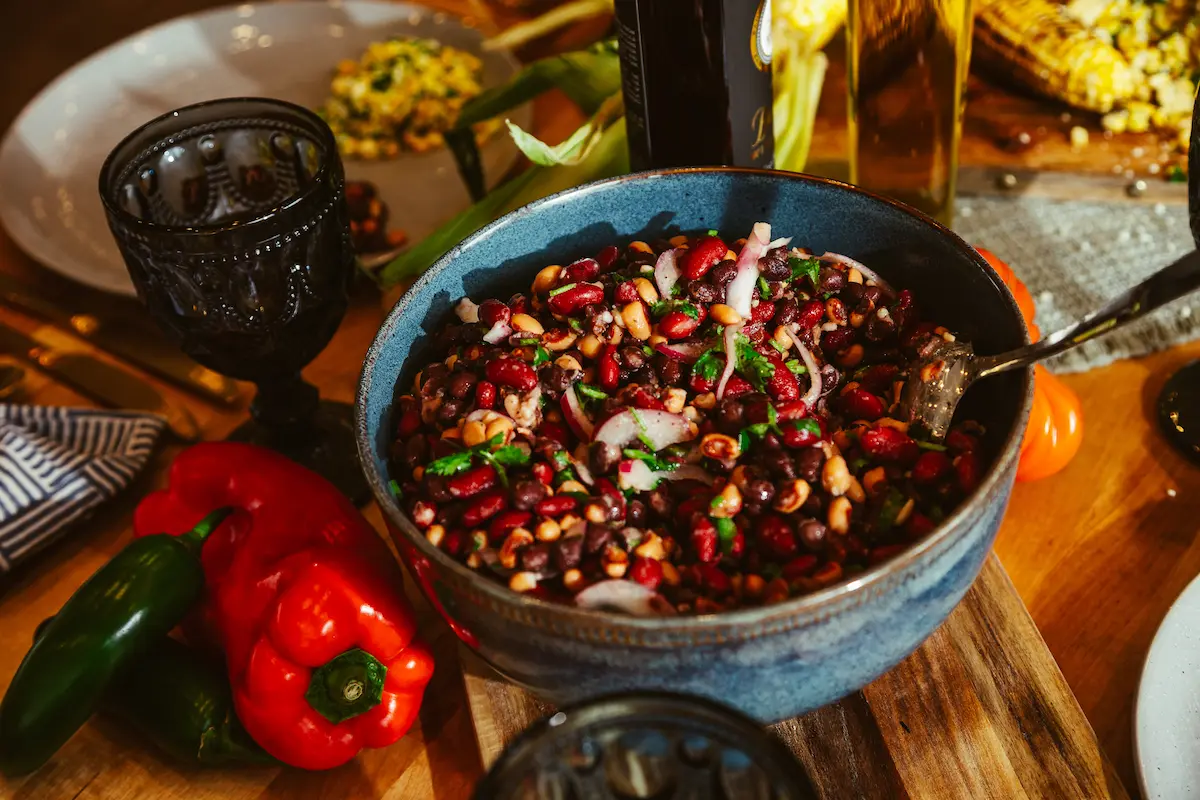 Bowl filled with mixed bean salad on a wooden board next to red and green peppers and textured glassware.