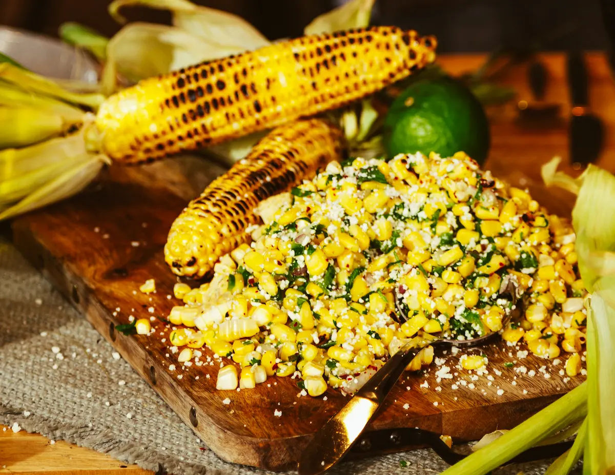 Wooden cutting board with grilled corn on the cob and a pile of corn salad mixed with herbs and cheese, next to a lime and corn husks.