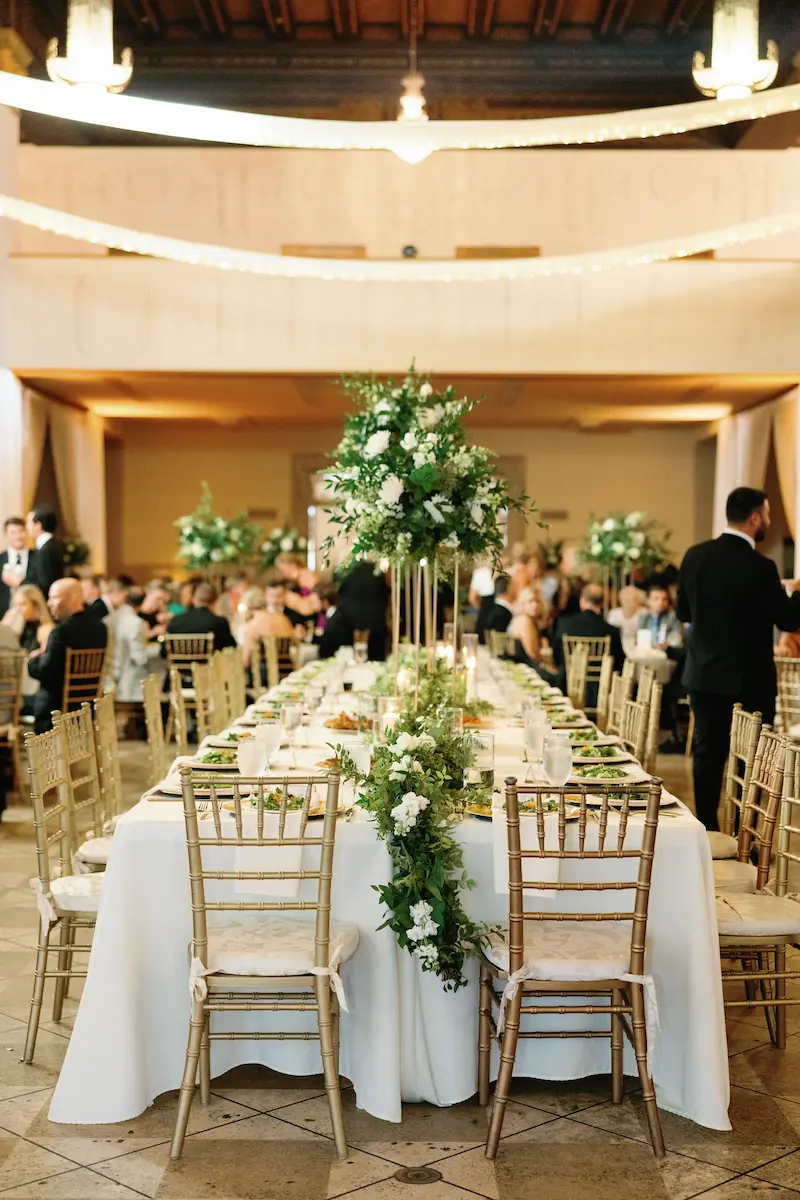 Elegant banquet hall with long table set for a formal event, decorated with tall white floral centerpieces and gold chairs.