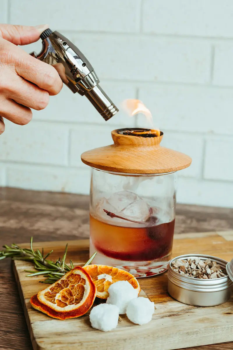 Hand using a torch lighter to ignite a flame on a wooden lid covering a glass with a cocktail and ice, on a wooden board with dried orange slices, cotton balls, rosemary sprigs, and a small metal container with dried herbs.