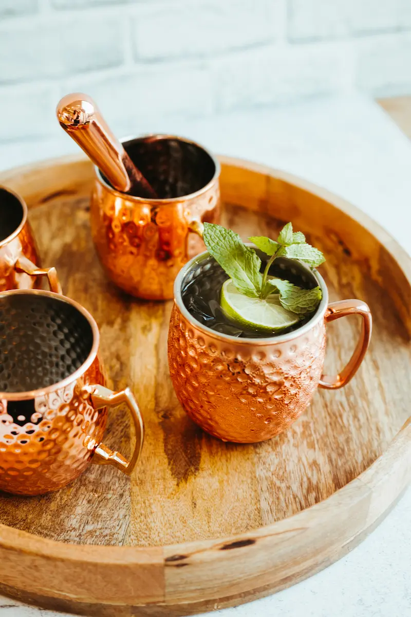 Copper mugs on a wooden tray, one filled with a drink garnished with a lime slice and mint leaves.
