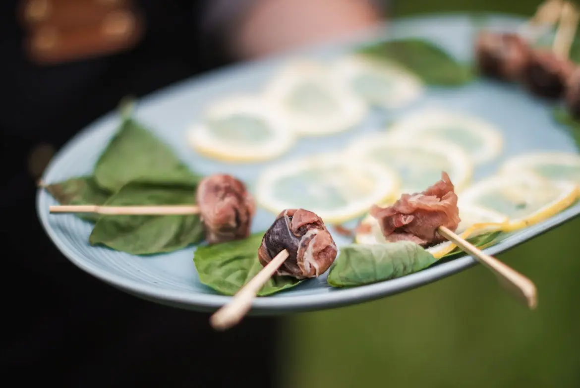 Blue plate with fresh green leaves and skewered pieces of cured meat, with lemon slices in the background.