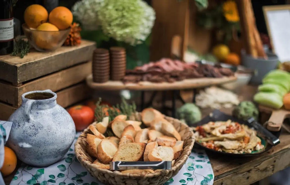 Table with basket of sliced bread, jar, bowl of oranges, and various dishes in a rustic setting.