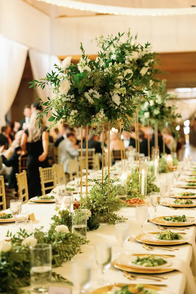 Long banquet table with white tablecloth, gold-rimmed plates, greenery garlands, glassware, and tall floral centerpieces at a formal event.