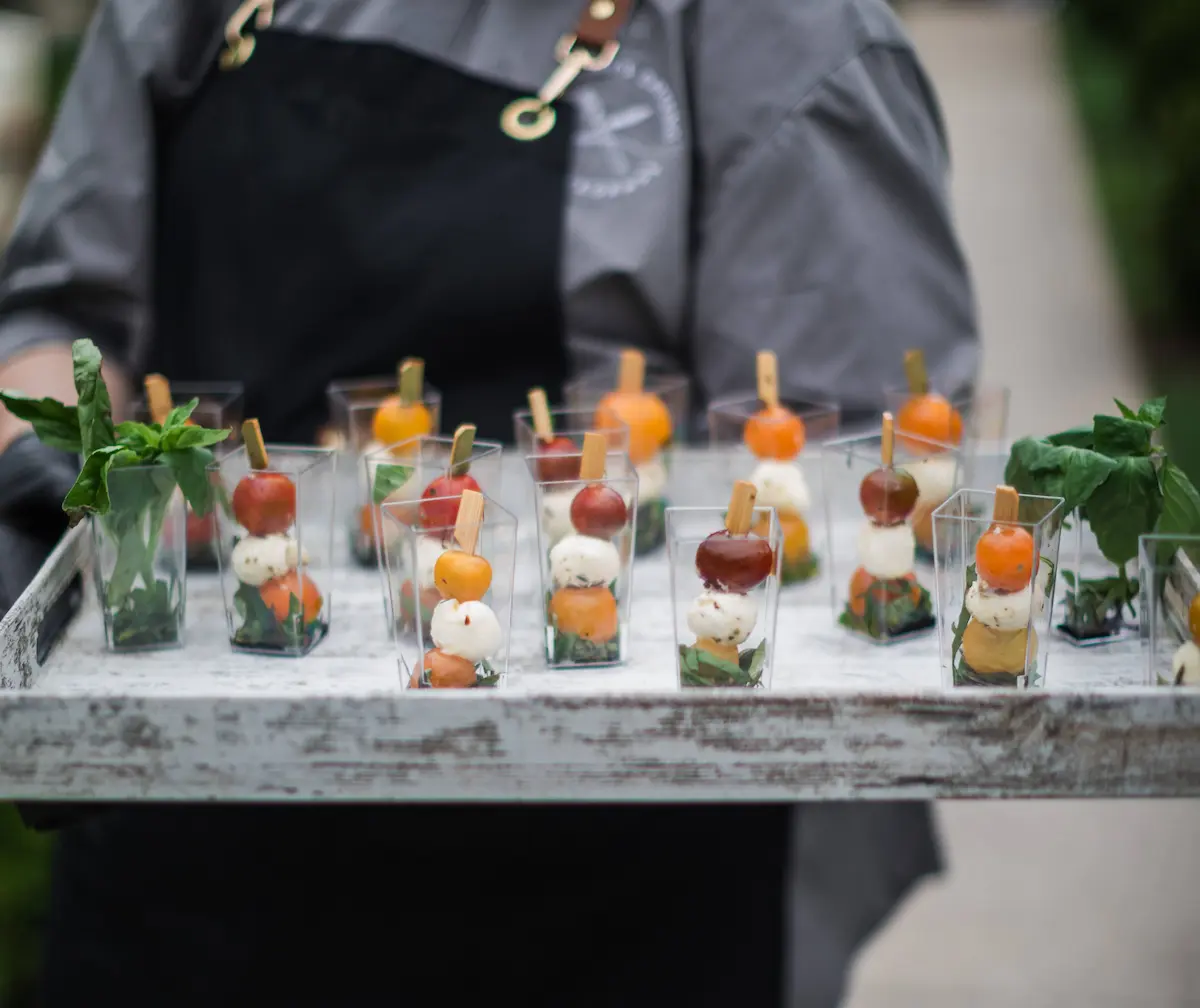 Tray of small cups with skewers holding cherry tomatoes, mozzarella balls, and basil leaves