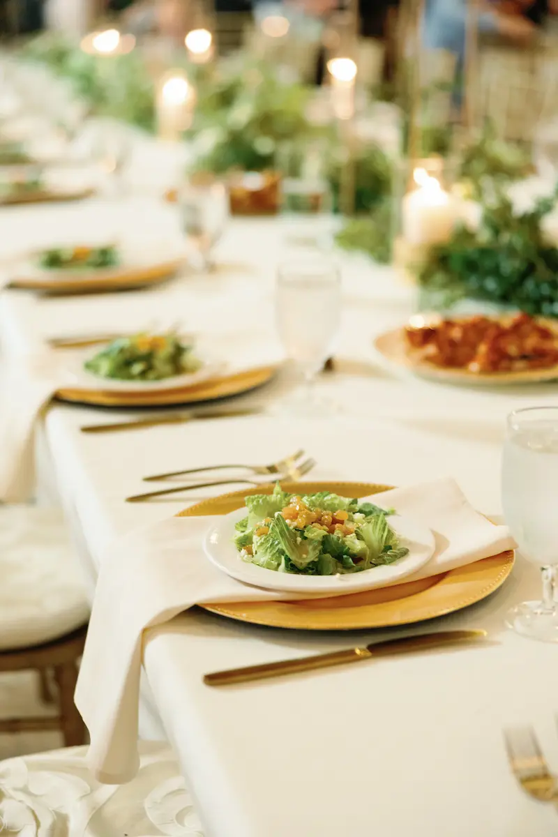 Elegant table setting with a fresh salad on a white plate over a gold charger, white napkin, gold flatware, and water glasses on a white tablecloth with blurred greenery and candles in the background.
