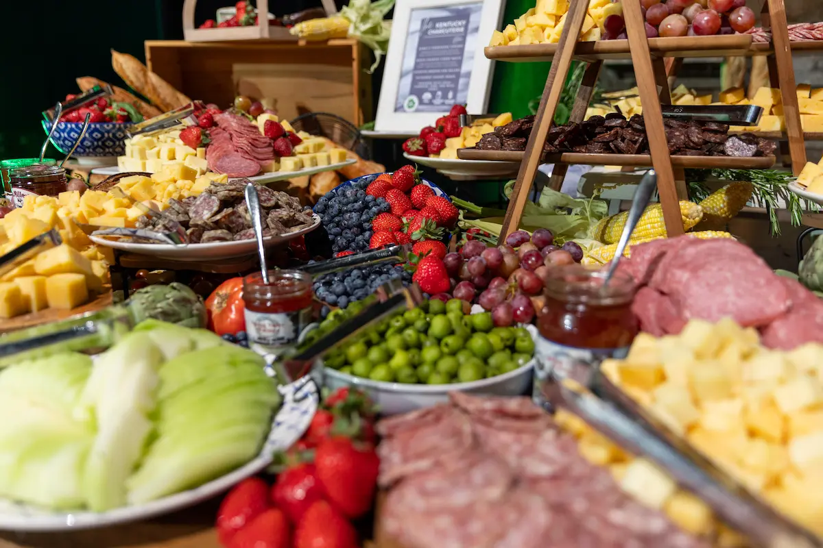 A colorful spread of assorted cheeses, sliced meats, fruit, green olives, artichokes, corn, and crackers arranged on platters and tiered wooden stands.