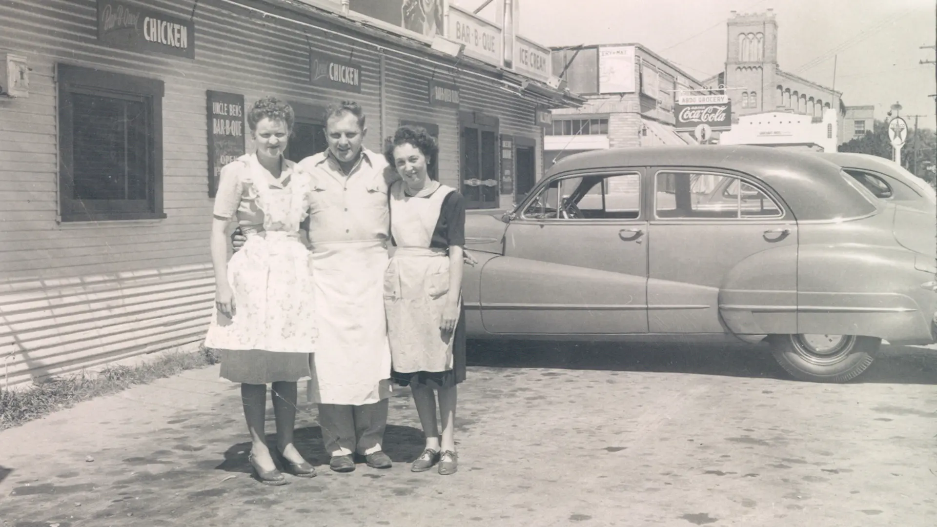 Black and white photo of two women and a man in aprons standing outside a restaurant with vintage cars parked nearby.